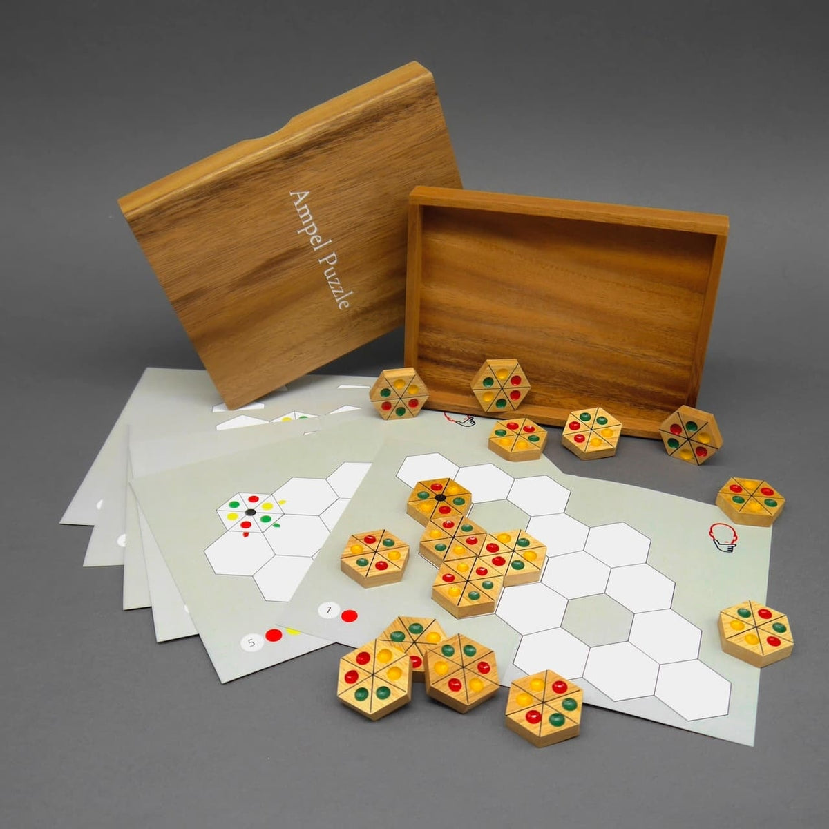Knobelspiel-Set: Offene Holzbox, mehrere Vorlagekarten und bunte hexagonale Spielsteine des Ampel Puzzle liegen auf grauem Hintergrund.