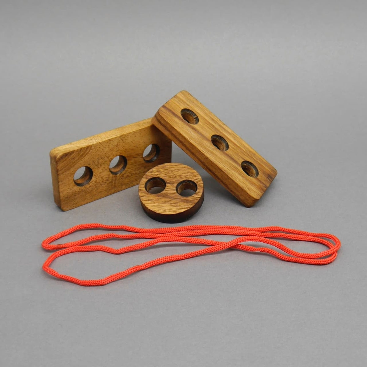 Knotenpuzzle: Zerlegtes Knotenpuzzle aus drei gelochten Holzformen – zwei rechteckige und eine runde – sowie einer roten Schnur vor neutralem Grau.