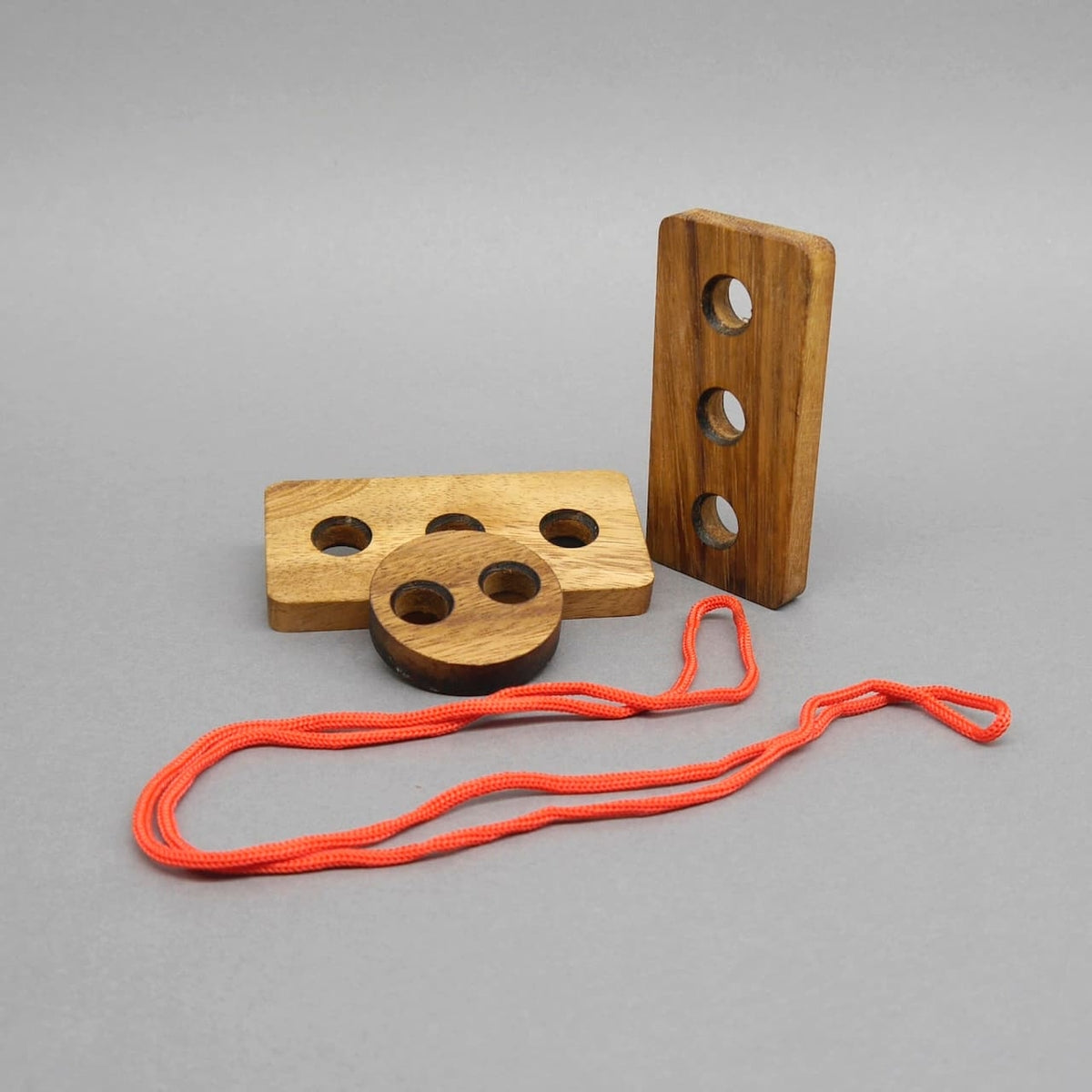 Logikpuzzle: Logikpuzzle für Kinder: rechteckiger Holzblock, stehende Holzleiste und runde Scheibe mit Lochungen, daneben lose rote Schnur.