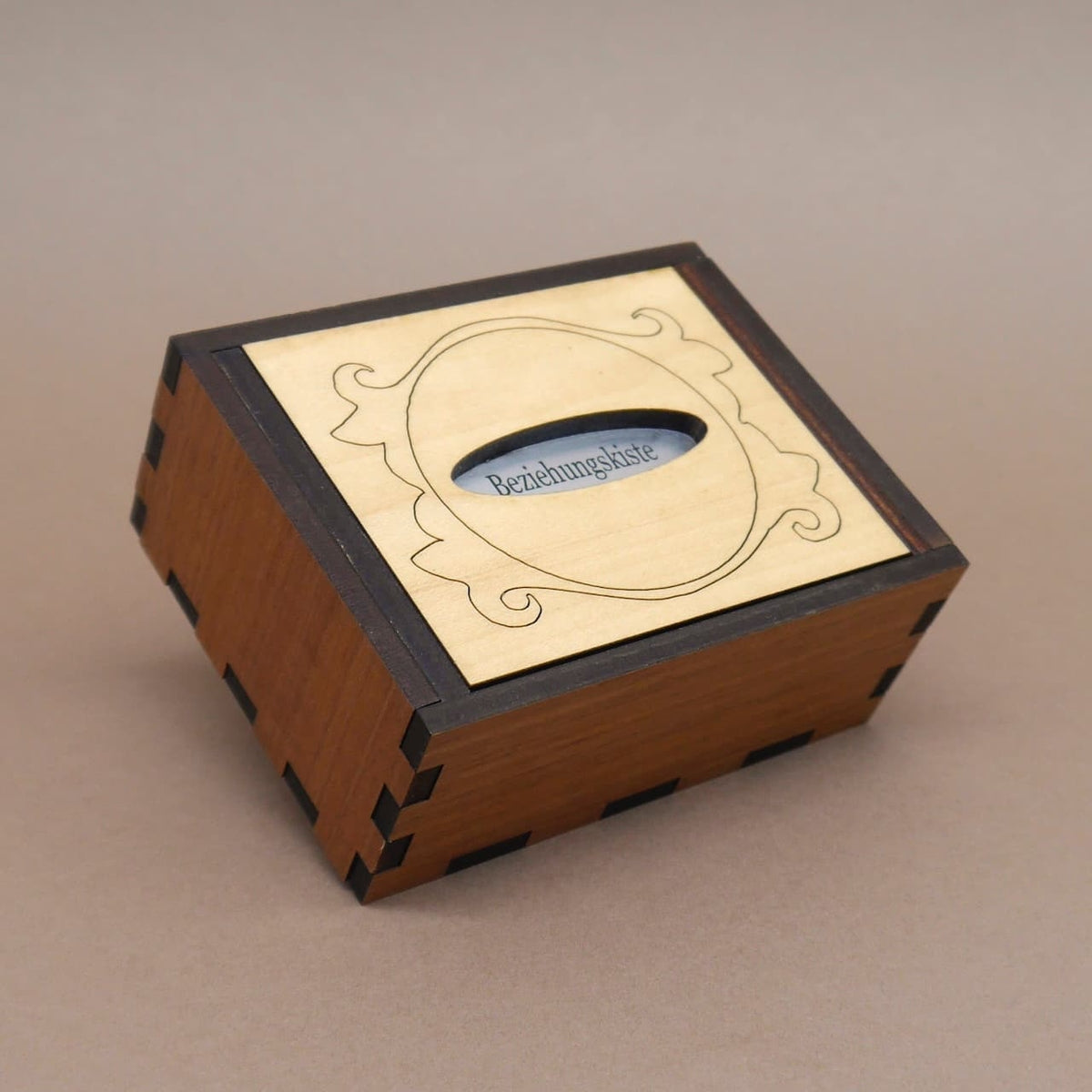 Geschlossene Puzzle‑Geschenkbox „Beziehungskiste“ aus dunklem Holz, diagonal aufgenommen; heller Deckel mit Ornamentrahmen.