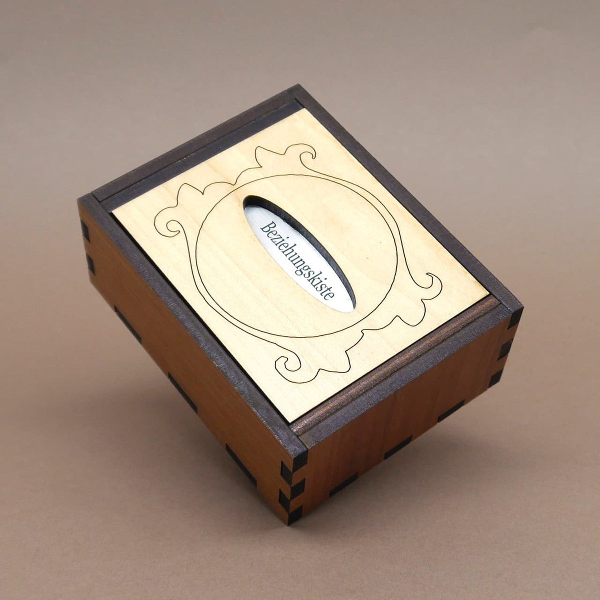 Trickbox „Beziehungskiste“ aus zweifarbigem Holz, geschlossen und nach hinten geneigt; heller Deckel zeigt Ornamentrahmen und Schlitz.
