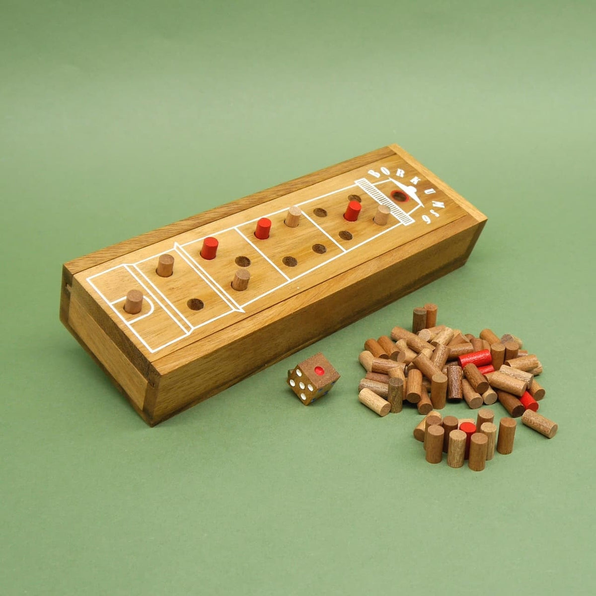 Beim detailreichen Kinderspiel „Borkums 6“ zeigt das längliche Holzbrett mehrere Spielfelder mit roten und naturfarbenen Stiften; daneben liegen Würfel und ein Haufen Holzsteine auf grünem Filz.