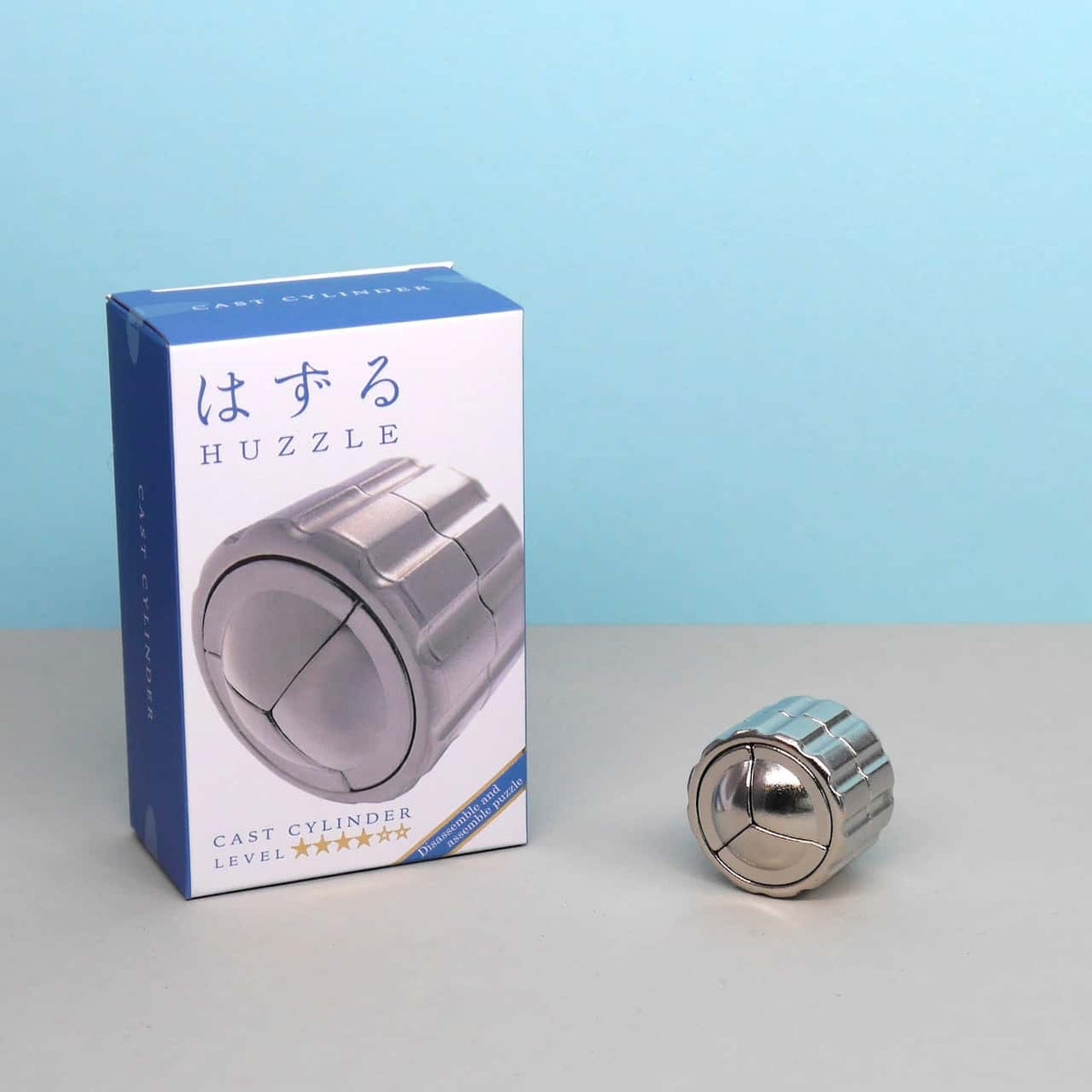 Metallisches Cast Cylinder Puzzle neben der Originalverpackung auf einem hellblauen Hintergrund – ideal für jeden Denkprofi und Knobelfan.