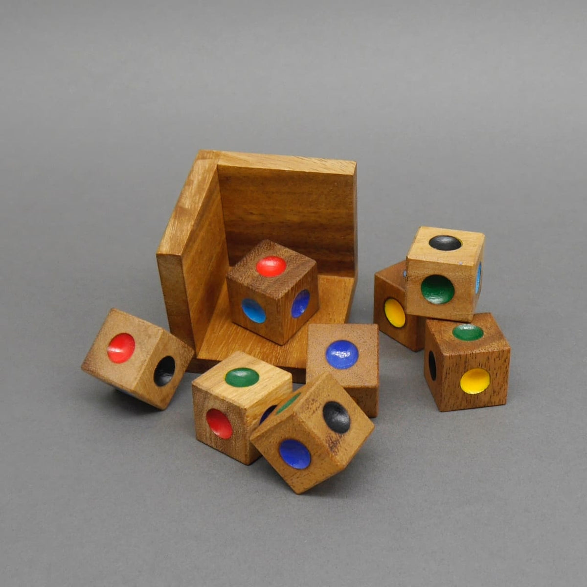 Zerlegtes Geduldspiel aus Holzwürfeln mit bunten Farbpunkten, verteilt vor einer offenen Holzbox auf grauem Untergrund.