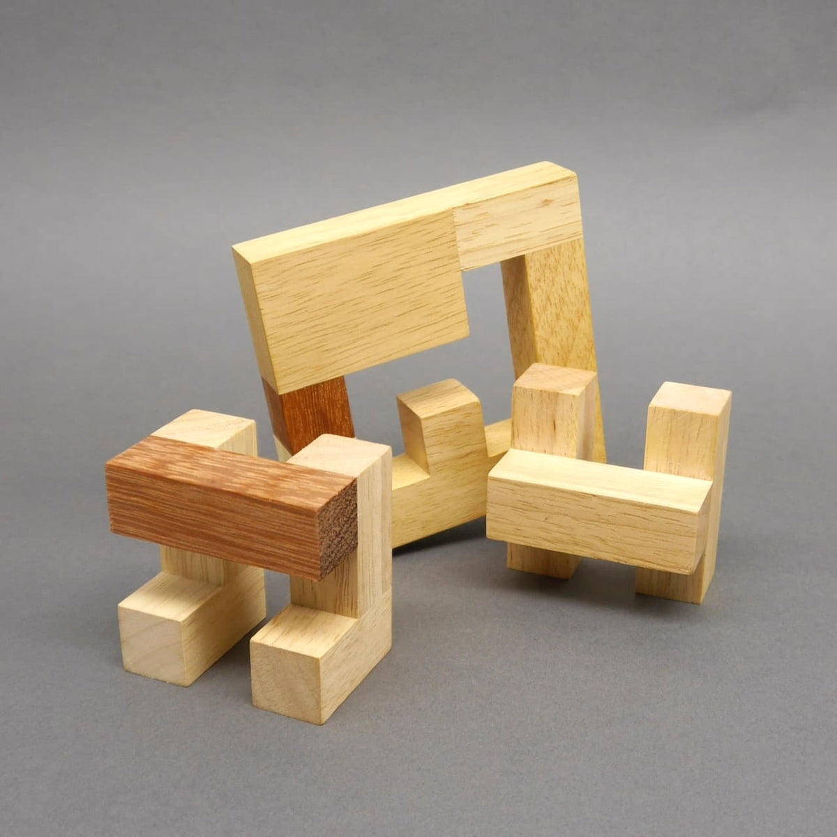 Mehrere Einzelteile eines hell‑dunkel gemaserten Holz‑Knobelspiel‑Puzzles sind locker vor einem grauen Hintergrund angeordnet.