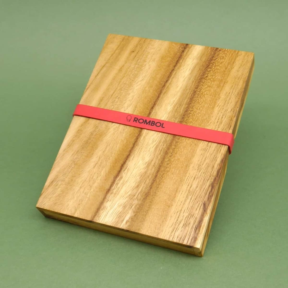 Geschlossene Holzpuzzle-Box aus hellem Holz, von einem roten Gummiband zusammengehalten, auf grünem Untergrund.