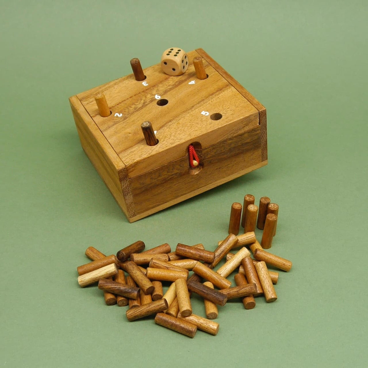 Familienspiel aus Holz: würfelförmige Box mit nummerierten Löchern, eingesteckten Stäbchen, Würfel und Holzstäbchenhaufen auf grünem Hintergrund.