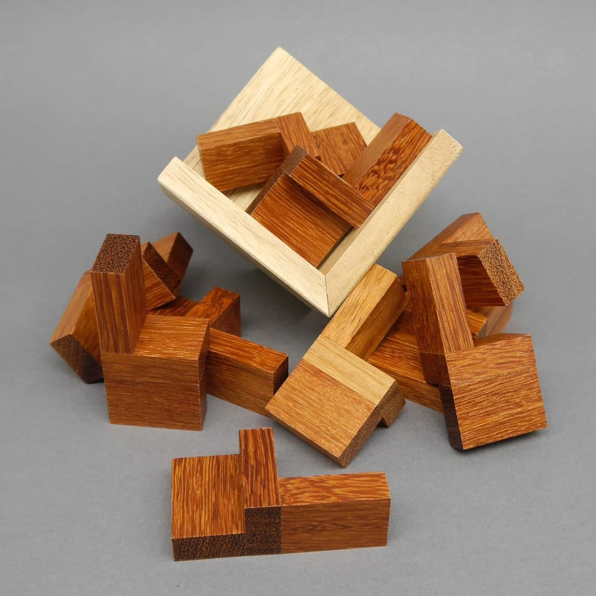 Zerlegte Holzstücke eines Brainteaser-3D-Puzzles liegen verstreut neben der hellen Rahmenbox auf grauem Hintergrund.