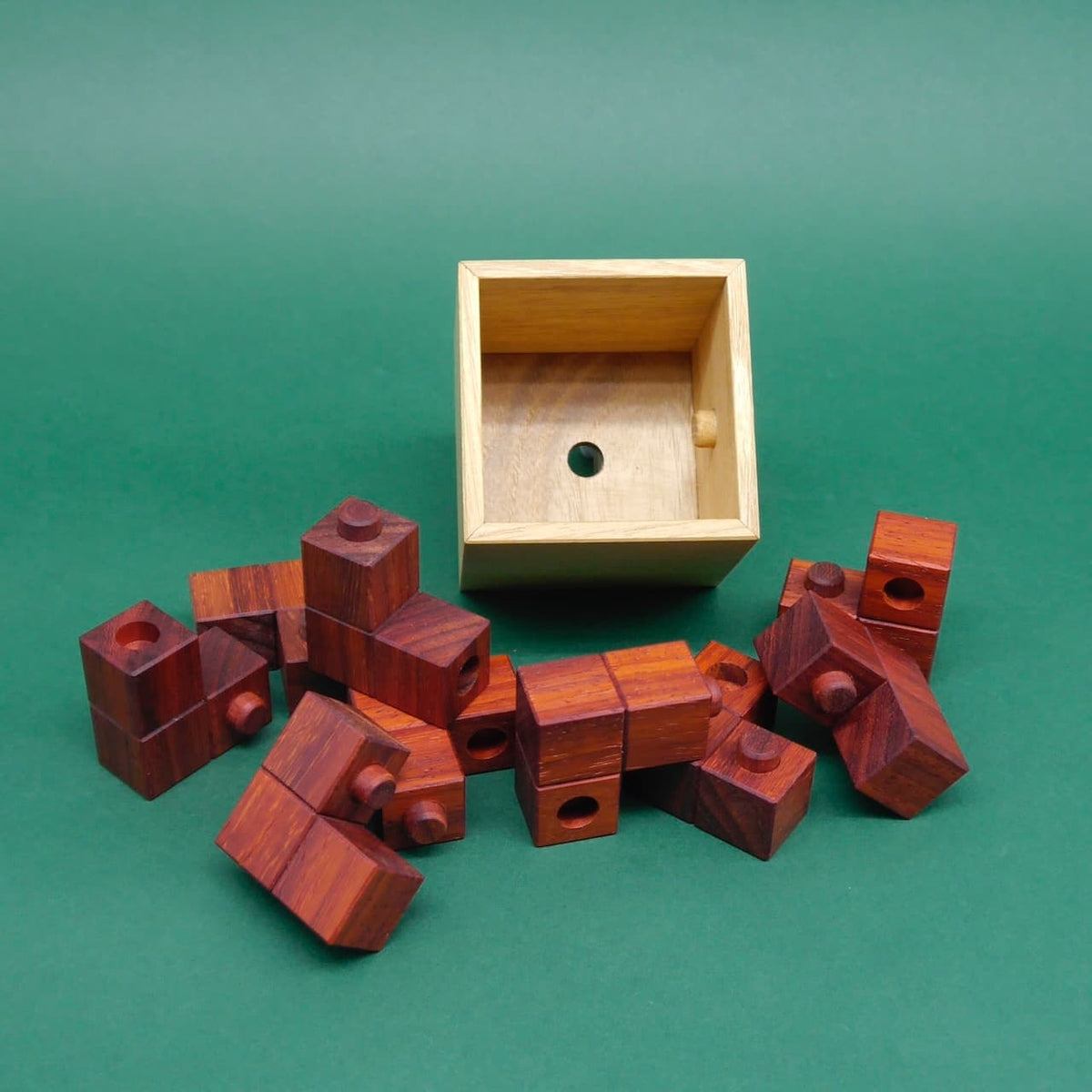 Rötlichbraune Würfelbausteine mit Zapfen und Bohrungen liegen verstreut vor einer leeren hellen Holzkiste – ein haptisches Denkspiel auf grünem Hintergrund.