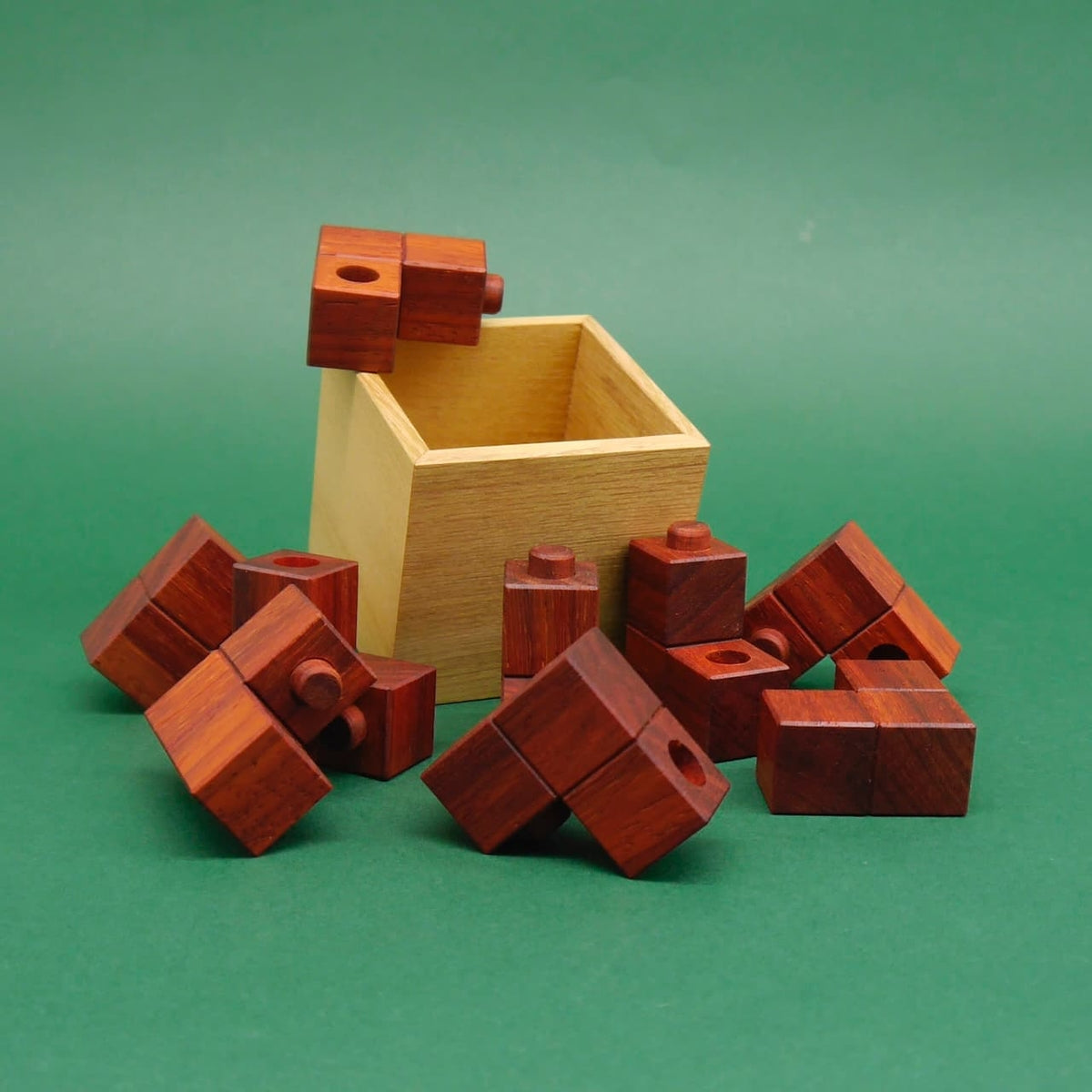 Ein aus Holz gefertigtes Geduldsspiel: Würfelbausteine umrunden eine offene Kiste, während ein Stein balanciert oben auf dem Rand.