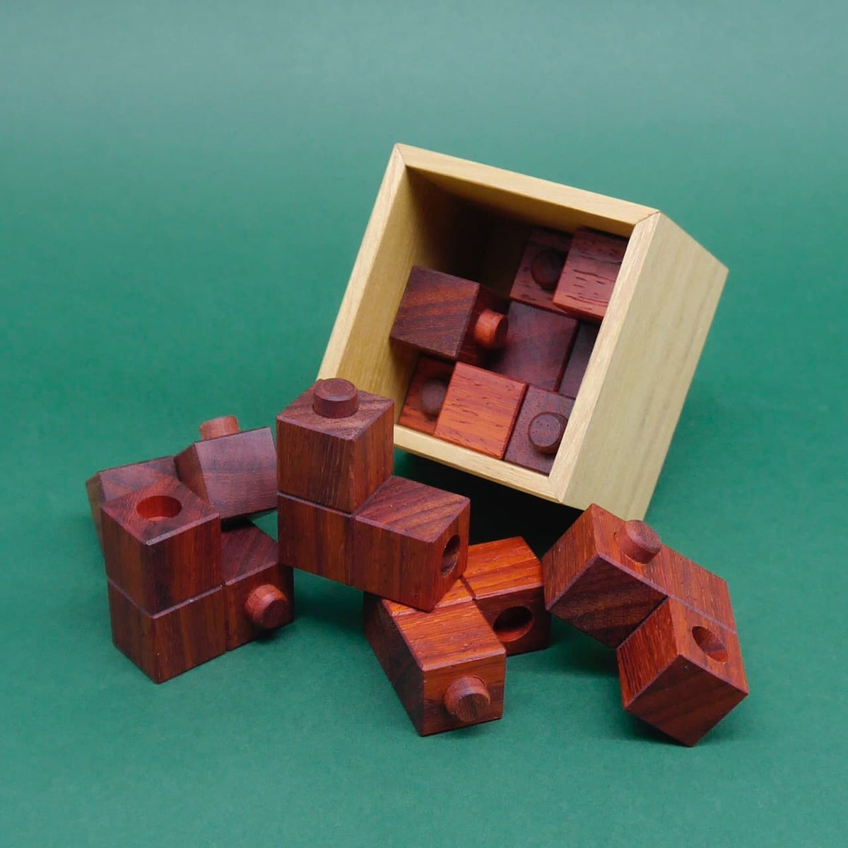 Aus einer gekippten Box purzeln mehrere ineinander gesteckte Holzwürfel‑Bausteine eines Holzpuzzles auf eine grüne Fläche.