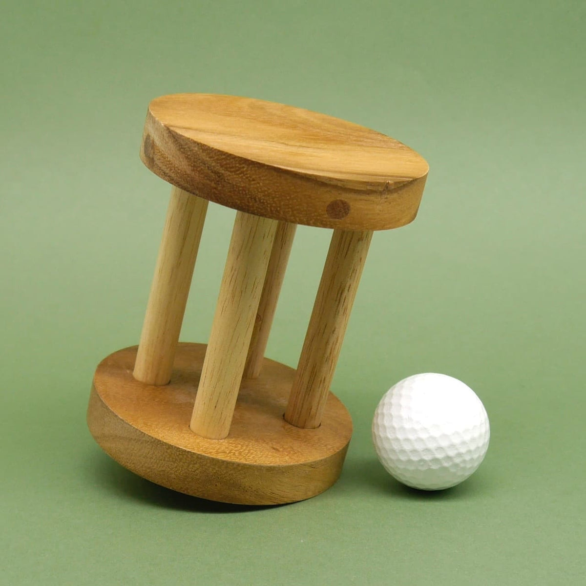 Kleines Holzpuzzle mit runder Basis lehnt seitlich, während ein loser weißer Golfball daneben auf dem grünen Untergrund liegt.