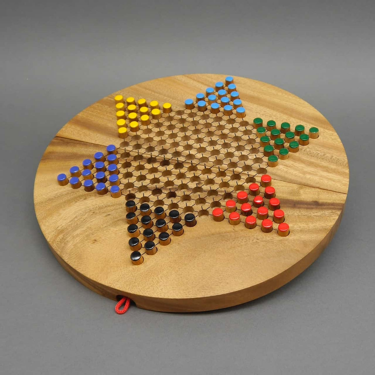 Ein rundes Halma-Brettspiel aus Holz in Sternform, mit farbigen Spielfiguren in Gelb, Blau, Schwarz, Rot und Grün, die in kleinen Löchern gesteckt sind. Das Spielbrett hat eine hochwertige Holzoptik und eine rote Schlaufe zum einfachen Verstauen.