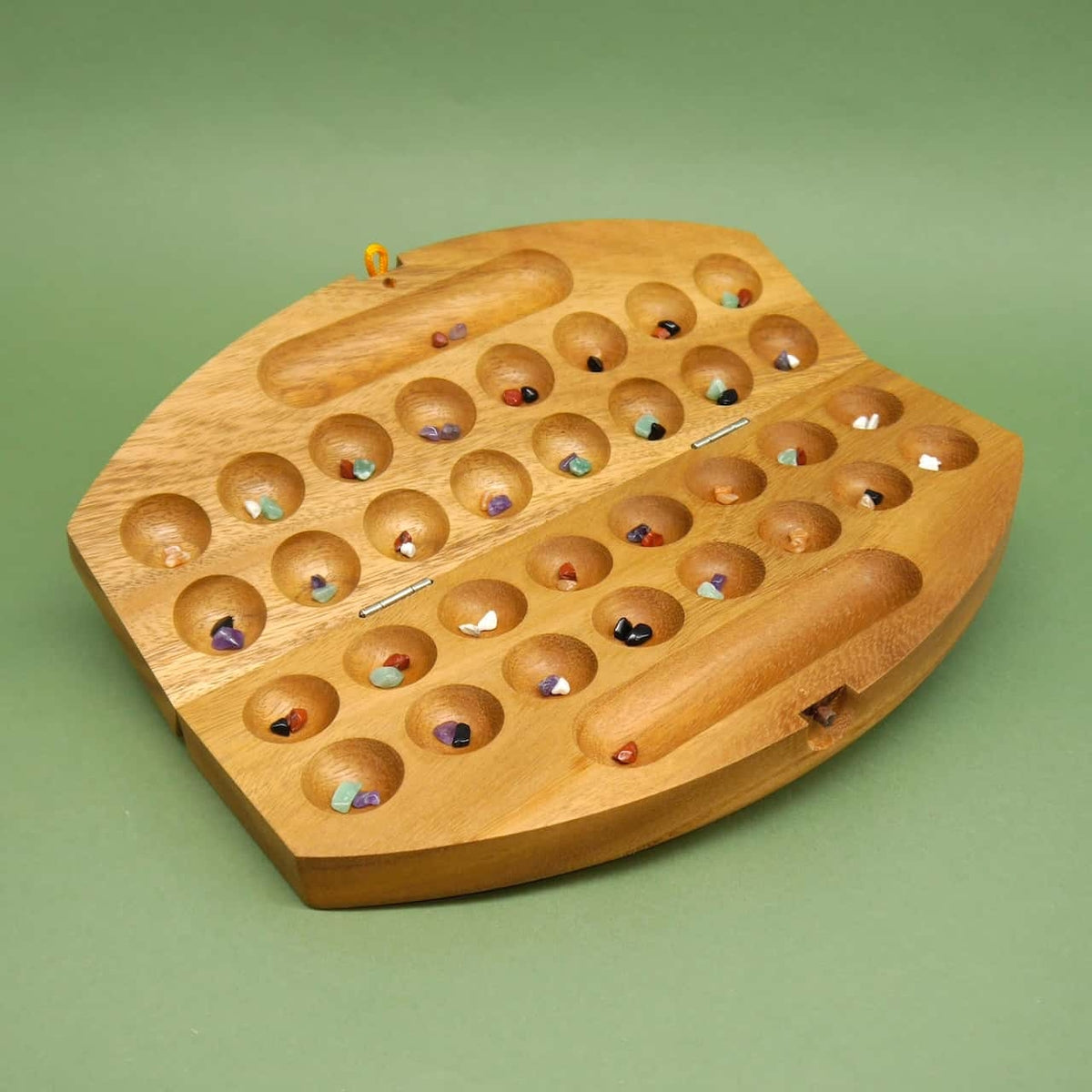 Geöffnetes Mancala-Holzspiel mit farbigen Steinchen in 32 runden Mulden auf grünem Untergrund.