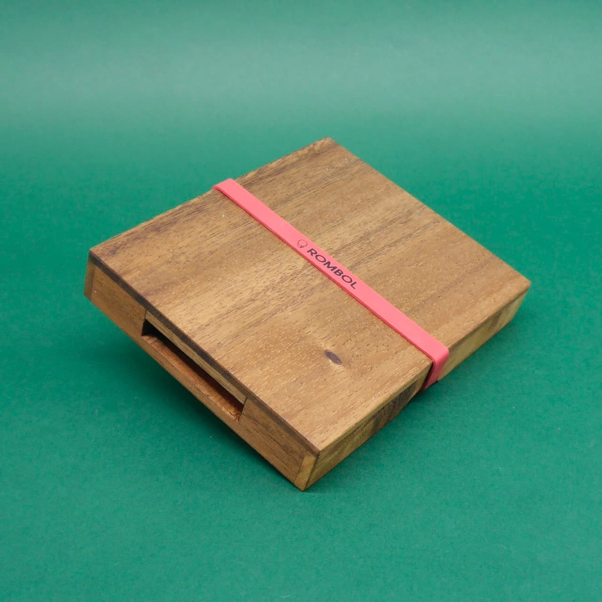 Geschlossenes Schiebepuzzle aus naturbelassenem Holz, von einem roten ROMBOL‑Gummiband gesichert, vor grünem Hintergrund.