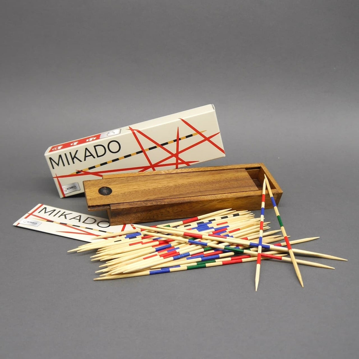 Komplettes Mikado-Set: offene Holzbox, Kartonverpackung und viele verstreute, bunt geringelte Holzstäbchen auf grauem Hintergrund.