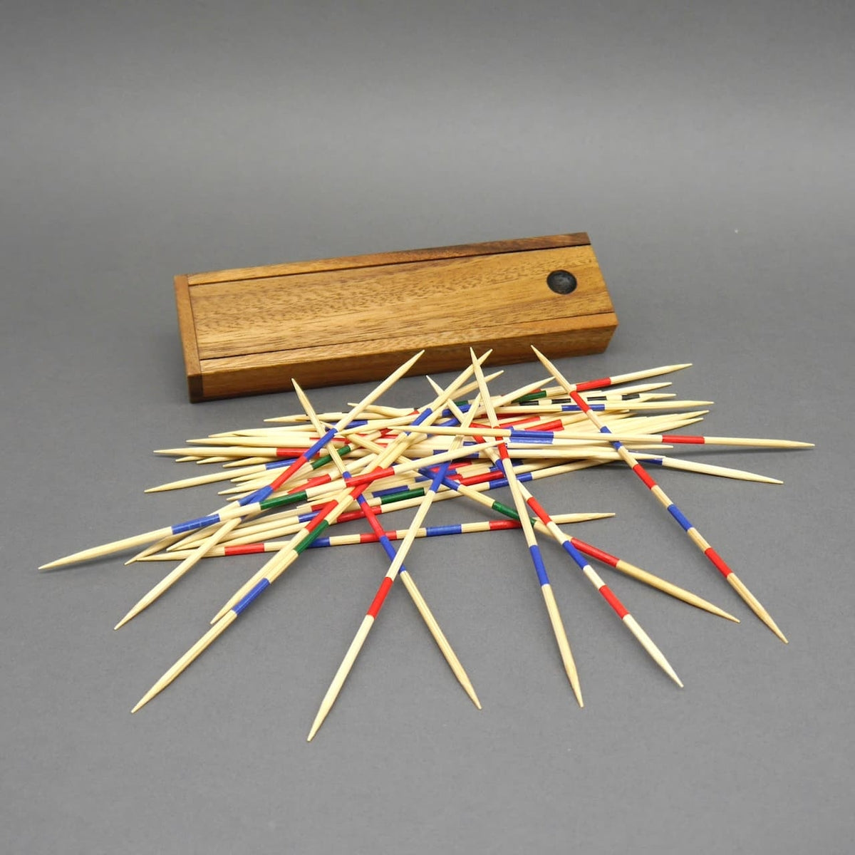 Dutzende Mikado-Stäbchen mit farbigen Markierungen liegen fächerförmig vor einer geschlossenen Holzbox auf grauem Untergrund.