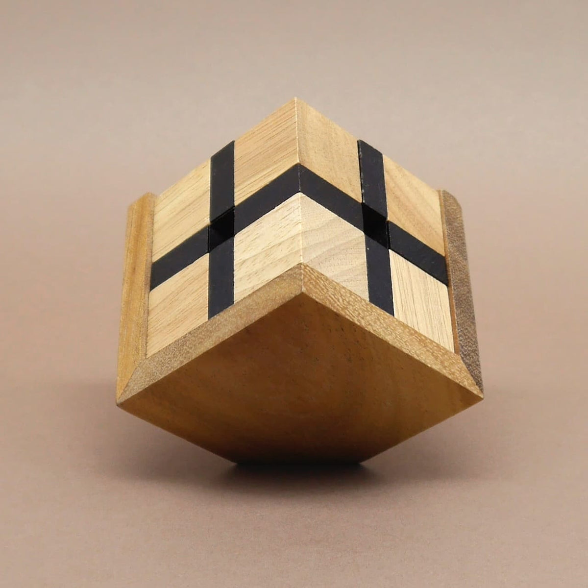 Holzpuzzle‑Würfel: heller Quader mit schwarzen Kreuzstreifen balanciert spitz auf einer Ecke vor neutralem Hintergrund.