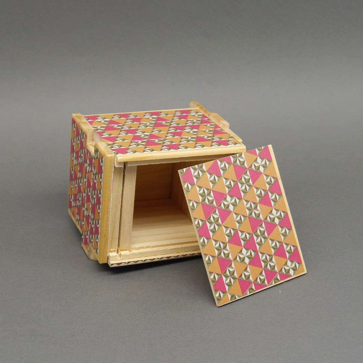 Geöffnete Holzpuzzle-Box im Osaka-Stil, zeigt das leere Innere und die abgenommene Frontabdeckung mit buntem Mosaikdesign.
