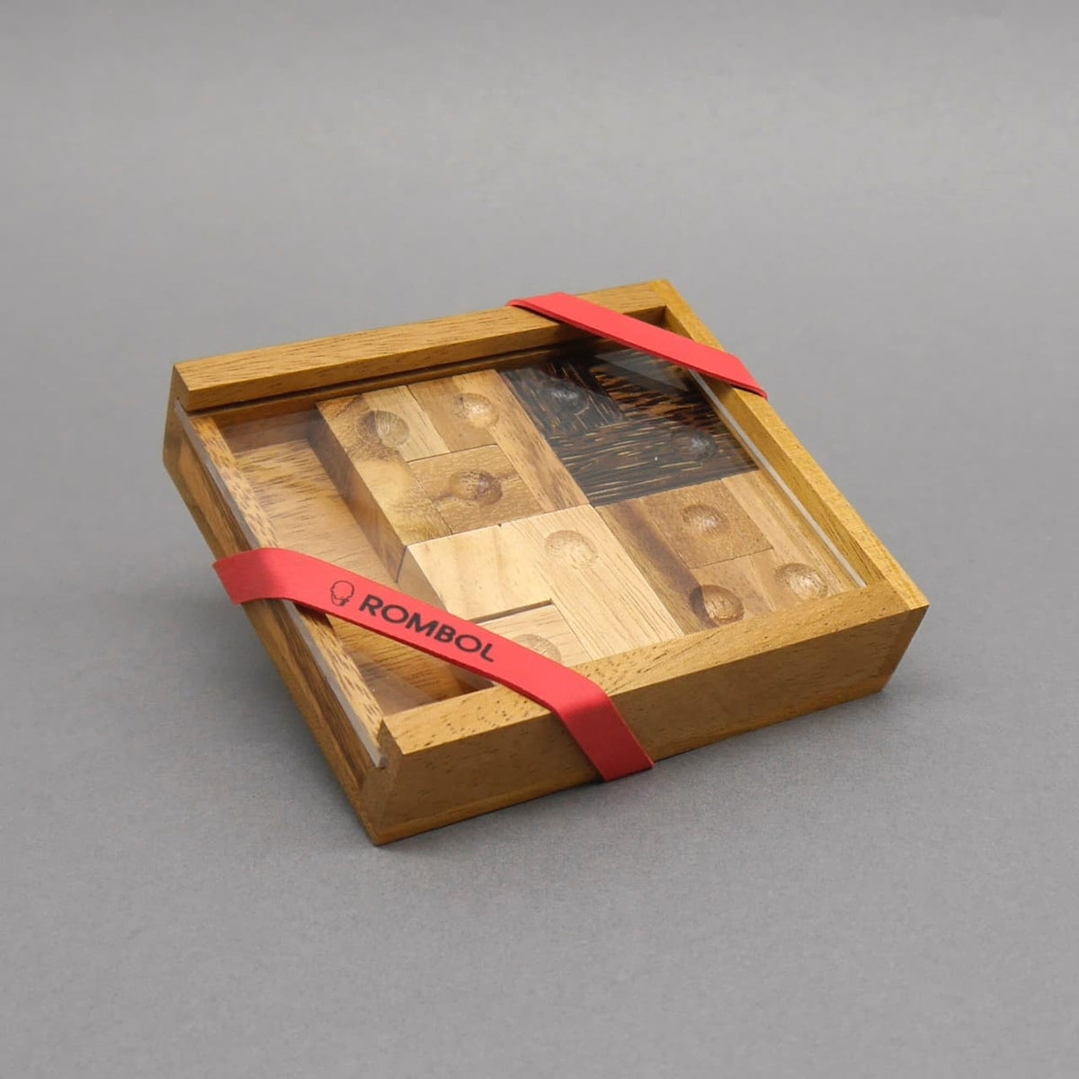 Geschenkverpacktes IQ‑Puzzle in Holzbox mit Klarsichtdeckel und rotem ROMBOL‑Band.