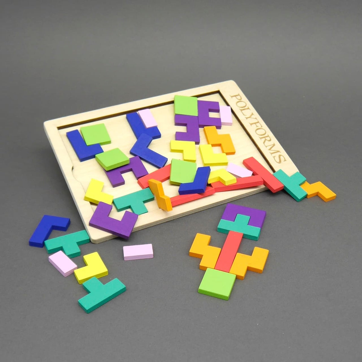 Holzrahmen mit dem Schriftzug „POLYFORMS“, in dem zahlreiche bunte geometrische Formen lose verteilt und teilweise aus dem Rahmen gefallen sind. Die Teile liegen auch auf der grauen Oberfläche davor.