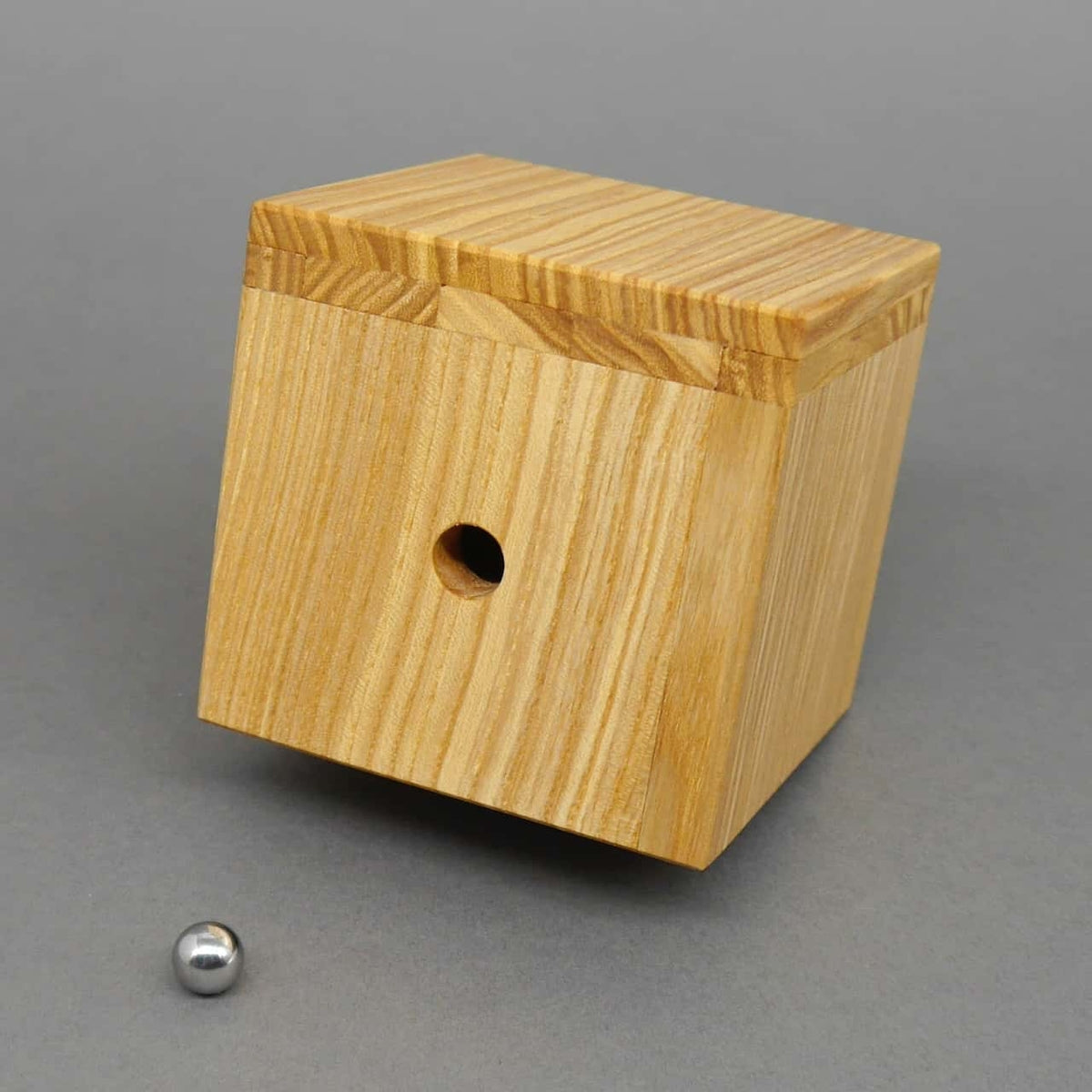 Holzpuzzle in Würfelform mit einem Loch in der Mitte und einer danebenliegenden Metallkugel auf grauem Hintergrund.