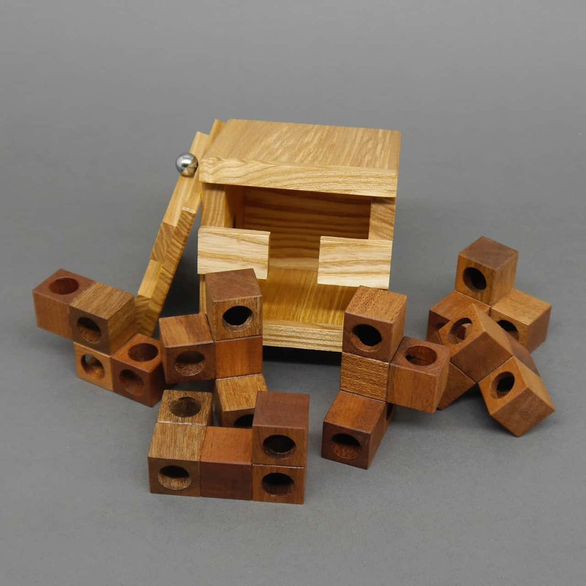 Geöffnete Holz-Box mit herausgenommenen braunen Puzzle-Elementen mit Löchern – ein Denkspiel aus Holz geöffneter Holz-Würfel mit eingesetzten Puzzle-Elementen, daneben liegt der abgenommene Deckel mit Metallkugel.