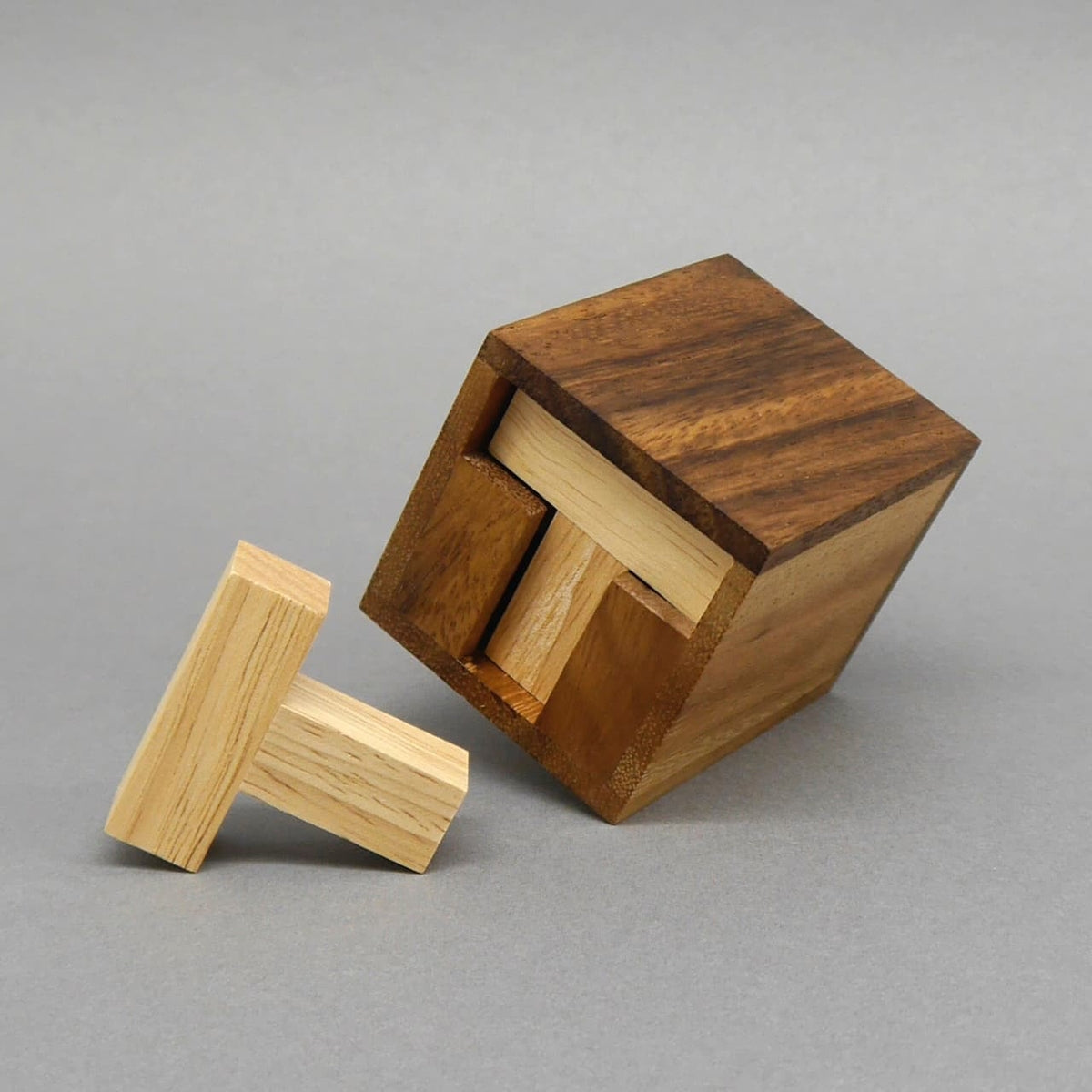 Braune Holzbox kippt auf den grauen Untergrund; zwei helle, T-förmige Holzsteine ragen heraus, einer liegt davor – zeigt das teilweise gelöste „Six T“-Packpuzzle.