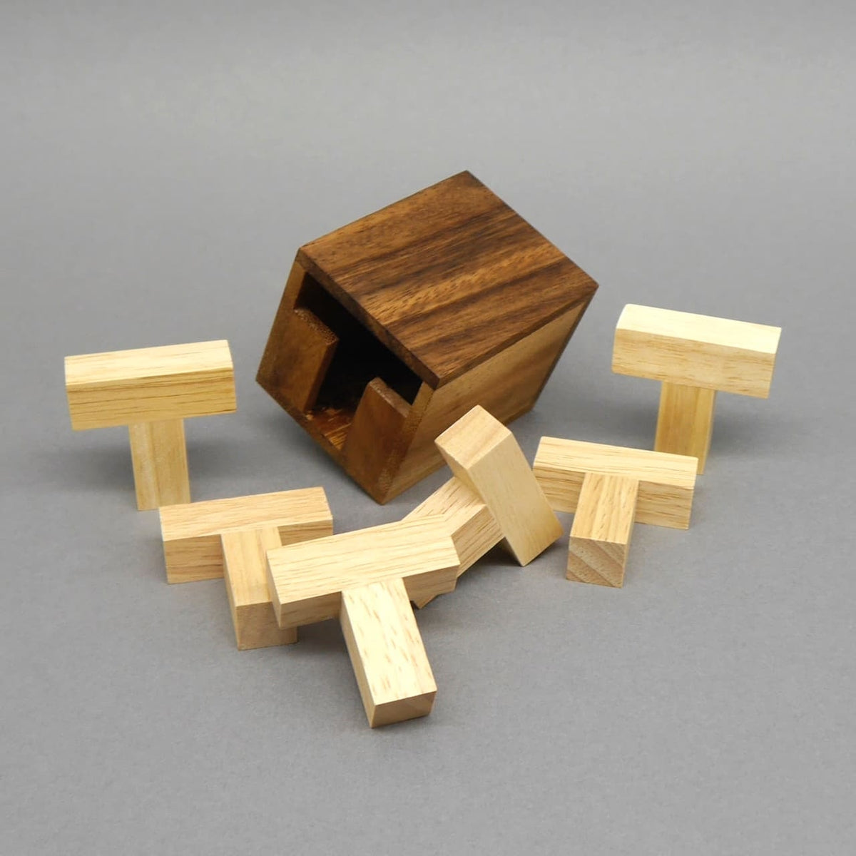 Geöffnete Holzbox neben allen sechs zerlegten, hellen T-Holzsteinen auf grauem Hintergrund – das „Six T“-Puzzle komplett auseinandergebaut.