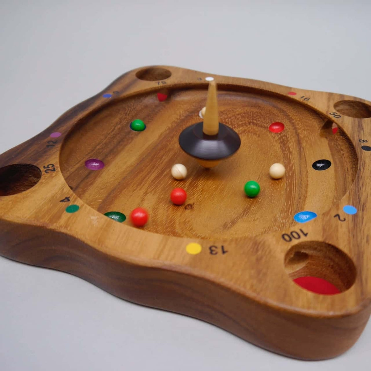 Schwungvolles Familienspiel „Tiroler Roulette“: Kreisel in Bewegung, verstreute Farbkugeln und nummerierte Felder auf einem geschwungenen Holzbrett.