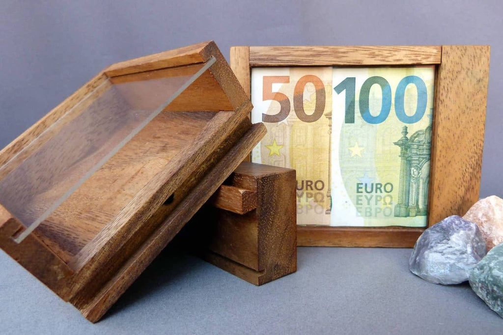 Holzbox mit Glasabdeckung, in dem ein 50-Euro- und ein 100-Euro-Schein nebeneinander platziert sind. Daneben liegen bunte Steine.