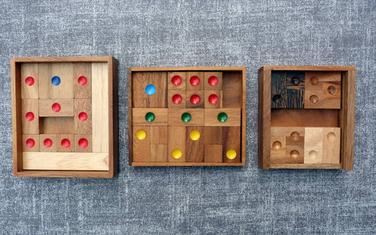 Drei hölzerne Schiebepuzzles nebeneinander auf einem grauen Stoffuntergrund, jeweils mit farbigen Markierungen in Rot, Blau, Grün und Gelb zur Unterscheidung der Teile.