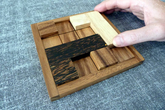 Hand bewegt ein Teil eines hölzernen Schiebepuzzles mit unterschiedlich gefärbten Holzstücken auf einem grauen Stoffuntergrund.