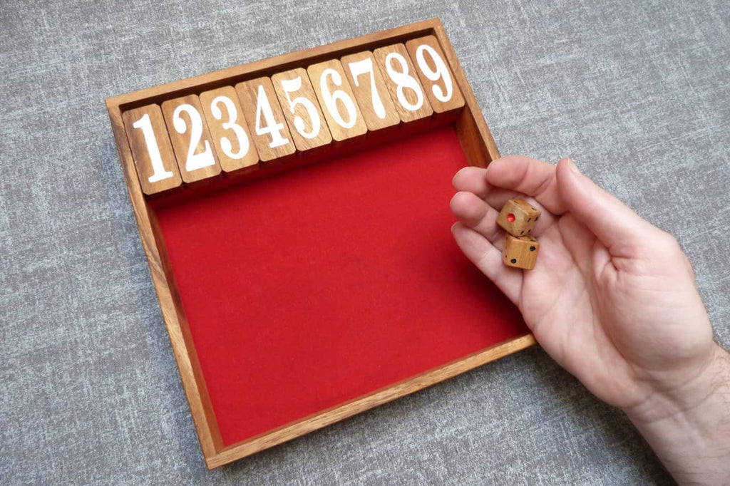 Holzspiel Shut the Box mit roten Filzeinsatz. Oben liegen die hochgeklappten Zahlenplättchen 1 bis 9, rechts hält eine Hand zwei kleine Holzwürfel.