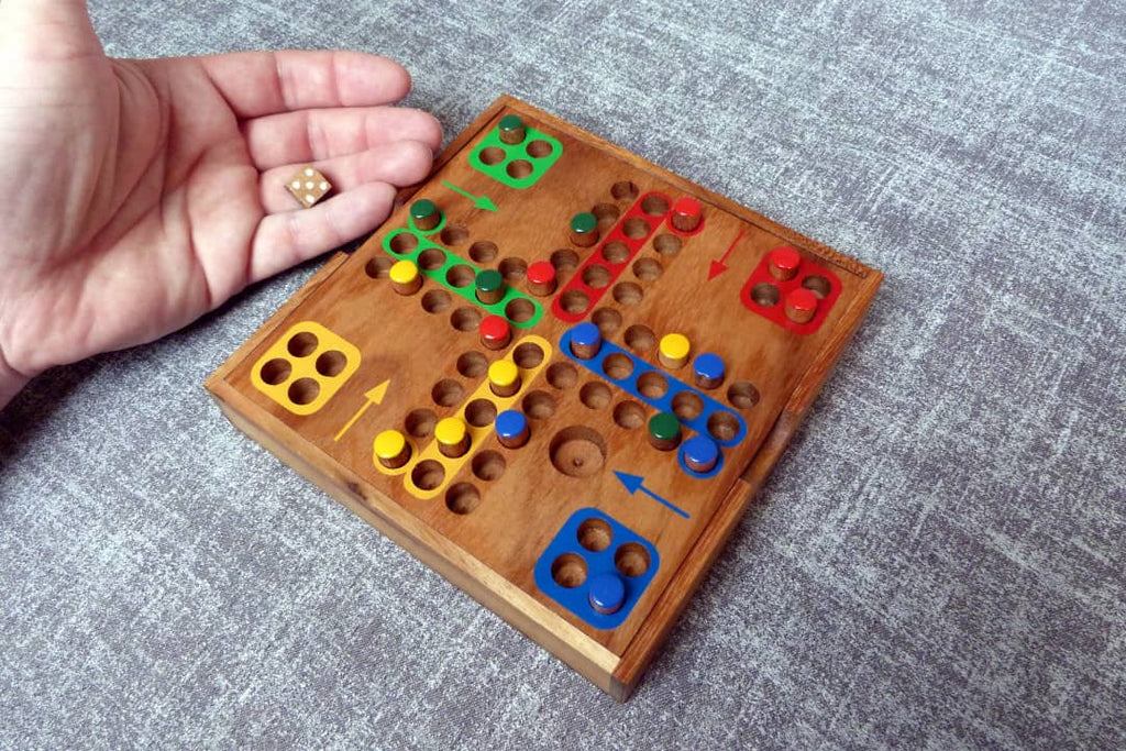 Eine Hand hält einen Würfel mit der Augenzahl vier über einem hölzernen „Mensch ärgere dich nicht“-Spielbrett. Auf dem Brett stehen bunte Spielfiguren in den Farben Rot, Gelb, Grün und Blau.