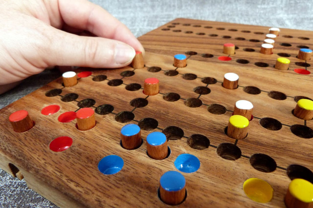 Eine Hand bewegt eine Spielfigur auf einem hölzernen Blockade-Spielbrett. Mehrfarbige runde Spielfiguren in Rot, Blau, Gelb und Weiß stecken in Vertiefungen des Brettes.