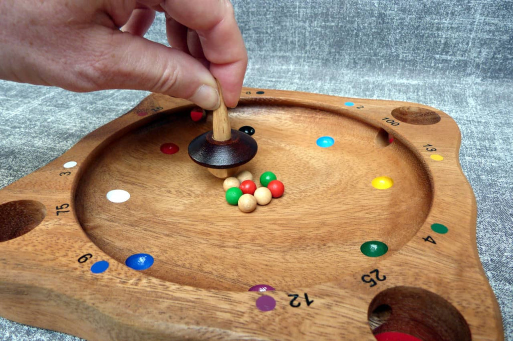 Hand dreht einen Kreisel auf einem runden Holzspielbrett mit bunten Kugeln und nummerierten Punkten.