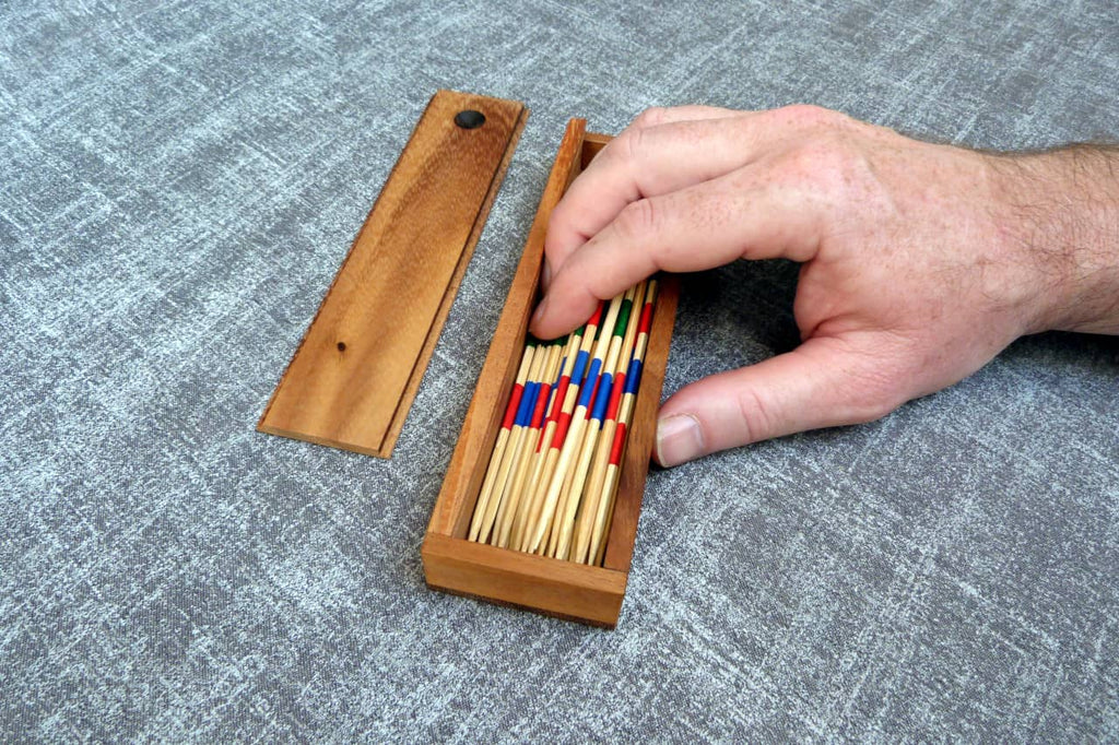 Eine Hand greift in eine hölzerne Schachtel mit Mikado-Stäbchen in verschiedenen Farben auf grauem Stoffuntergrund.