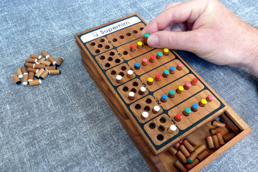 „Eine Hand steckt einen farbigen Spielstein in ein hölzernes Mastermind-Spielbrett mit der Aufschrift ‚Superhirn‘. Mehrere Reihen bunter Spielsteine sind bereits eingesetzt, daneben liegen weitere Holzsteine.