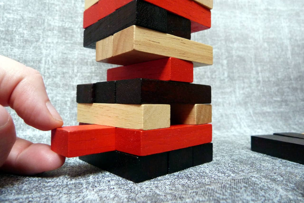 Nahaufnahme eines Jenga-Turms aus roten, schwarzen und naturfarbenen Holzklötzen. Eine Hand zieht vorsichtig einen roten Stein aus der unteren Reihe heraus.