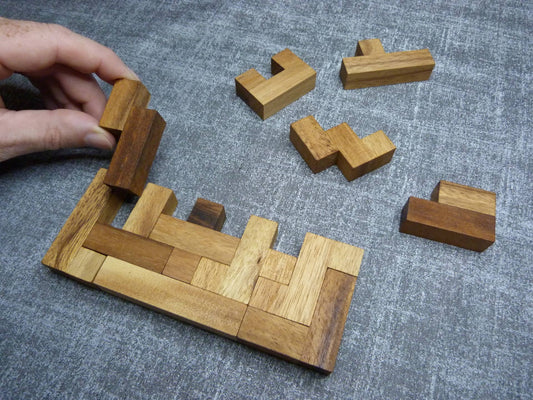 Eine Hand hält ein Holzpuzzle-Teil über ein teilweise zusammengesetztes rechteckiges Puzzle aus verschiedenen braunen Holzteilen, die auf einem grauen Stoffuntergrund liegen. Weitere Puzzleteile liegen ungeordnet daneben.