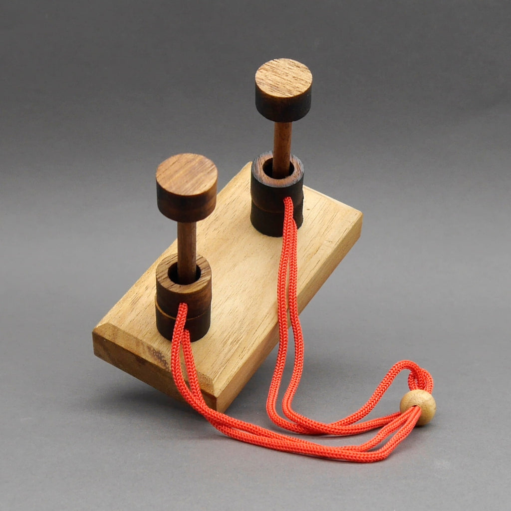 2 Pilone Seilpuzzle aus Holz auf schräg liegender Basis mit rotem Seil und Holzperle als Knotenpunkt.