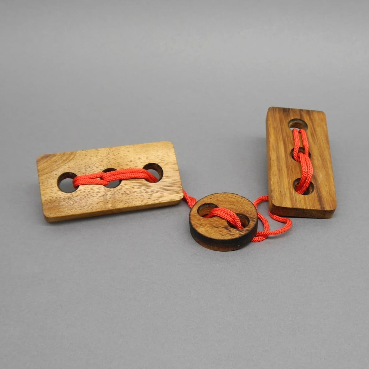 Holzpuzzle: Holzpuzzle mit zwei rechteckigen Holzklötzen und einer runden Scheibe, verbunden durch leuchtend rotes Kordelseil auf grauem Hintergrund.