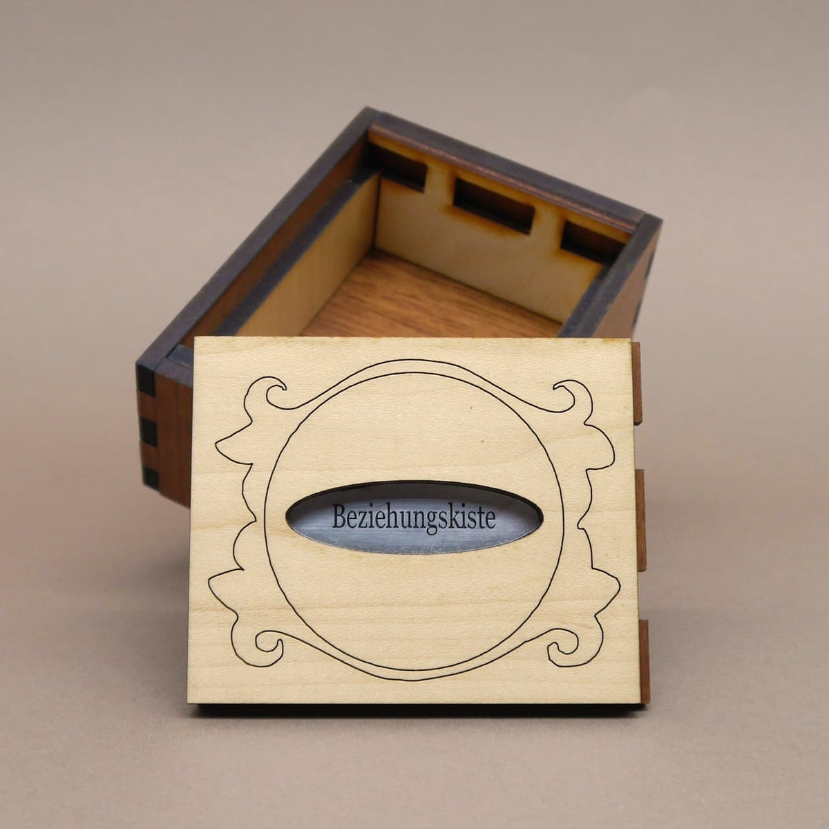 Geöffnete Holz‑Puzzlebox „Beziehungskiste“; hell gravierter Deckel mit ovalem Sichtfenster steht vor dunkelbraunem Korpus.