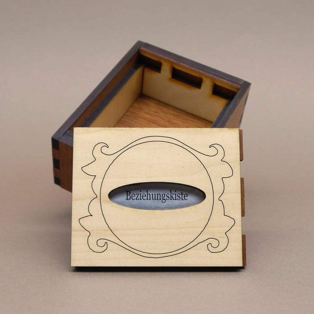 Geöffnete Holz‑Puzzlebox „Beziehungskiste“; hell gravierter Deckel mit ovalem Sichtfenster steht vor dunkelbraunem Korpus.