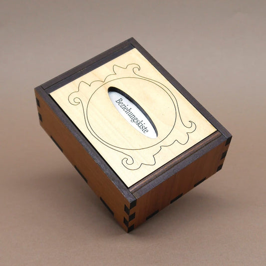 Trickbox „Beziehungskiste“ aus zweifarbigem Holz, geschlossen und nach hinten geneigt; heller Deckel zeigt Ornamentrahmen und Schlitz.