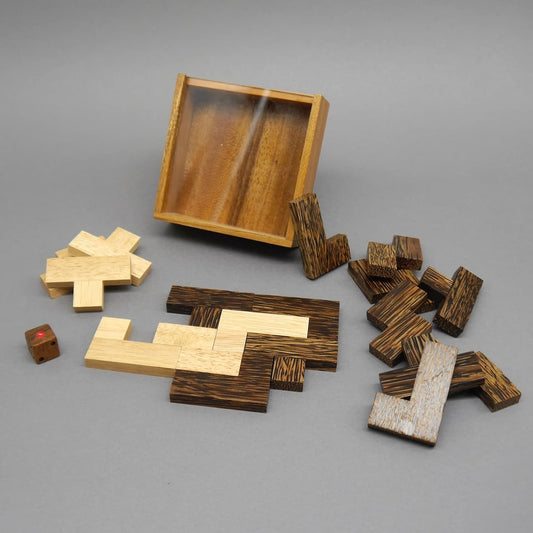 Offene Holzbox mit hell‑ und dunkelbraunen Puzzleteilen eines Brettspiel‑Sets sowie kleinem Würfel auf grauem Hintergrund.