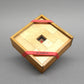 Quadratische Box, von roten Gummibändern gehalten, präsentiert ein kompaktes Holzspiel aus hellen Legeplättchen.