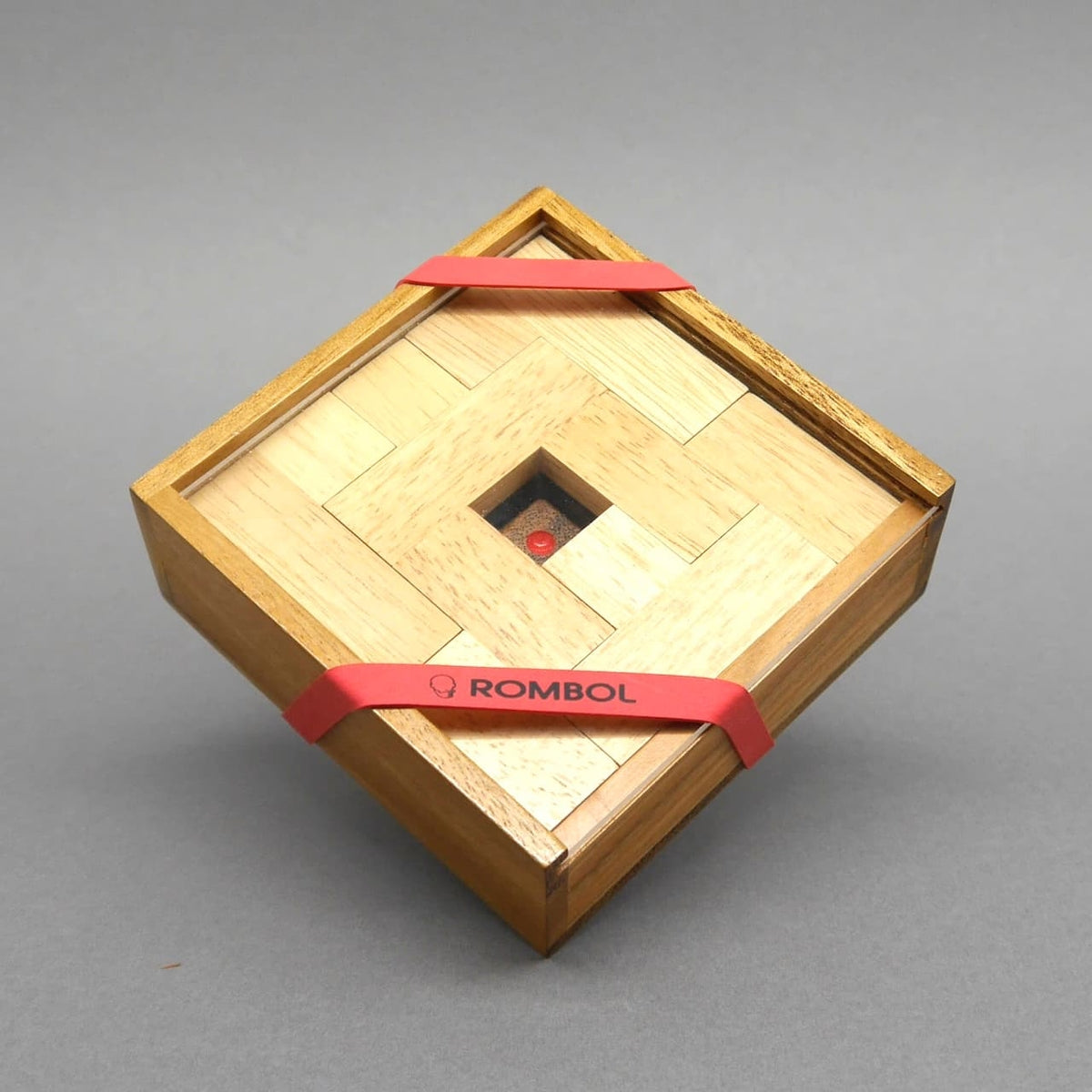 Quadratische Box, von roten Gummibändern gehalten, präsentiert ein kompaktes Holzspiel aus hellen Legeplättchen.