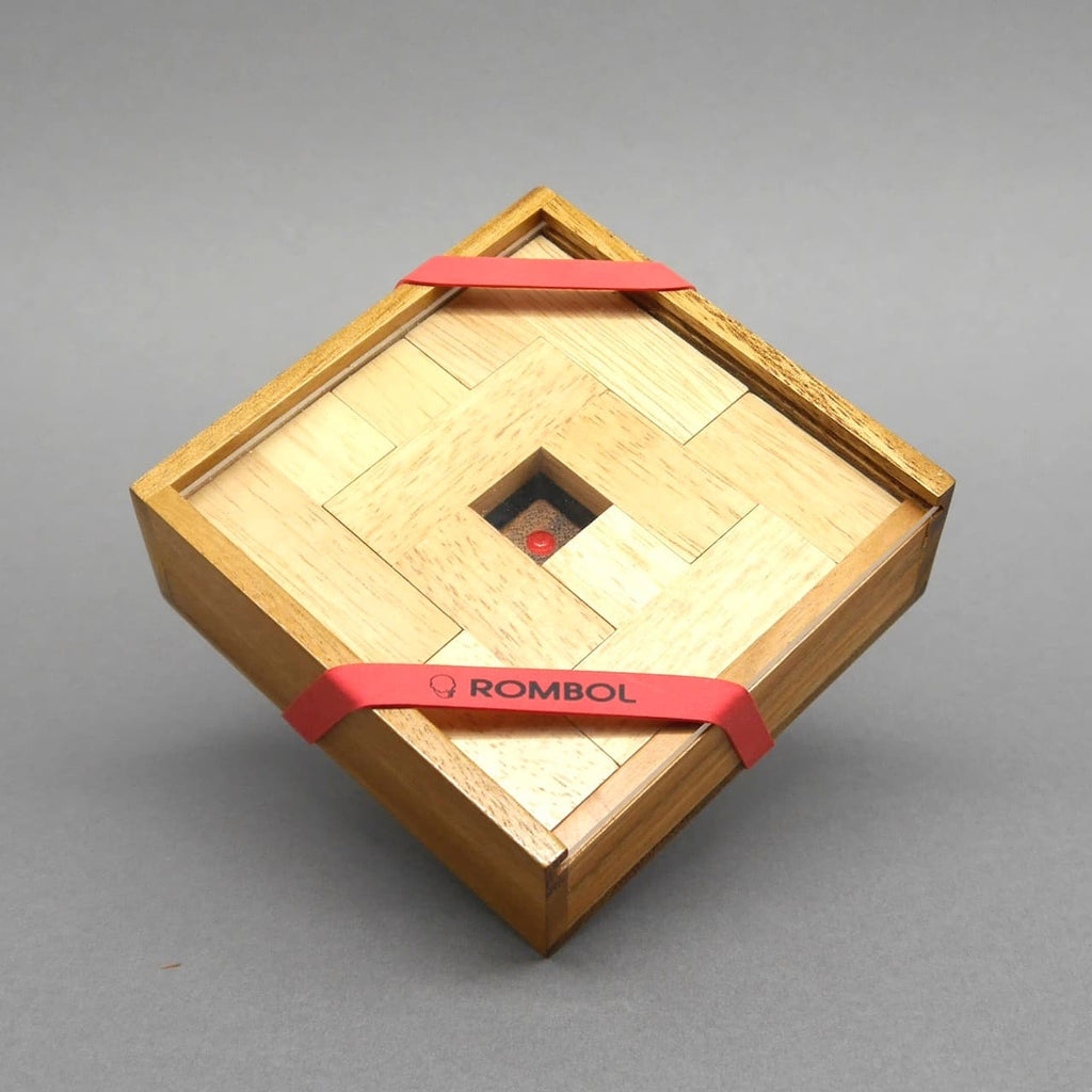 Quadratische Box, von roten Gummibändern gehalten, präsentiert ein kompaktes Holzspiel aus hellen Legeplättchen.